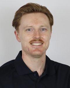 Portrait of a smiling man with red hair and fair skin in a black polo shirt.